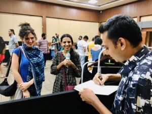 Photograph of the young writer Devasiachan Benny. He is signing an exObjects book at Bangalore Literature Festival, December 2024. Two young women smile as they watch.