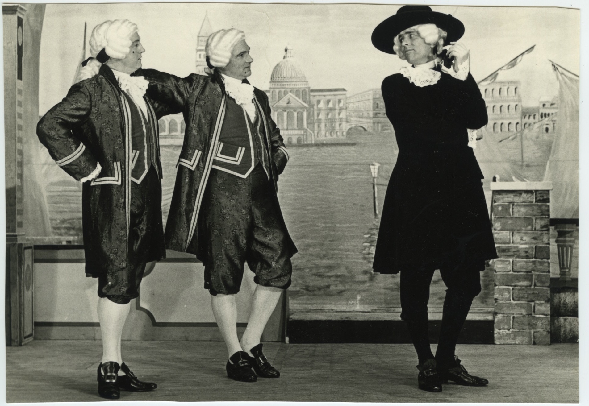 Three male actors in period costumes perform on a stage with a painted backdrop of the European city Venice, during a stage performance of ‘The Gondoliers’ by Gilbert and Sullivan.