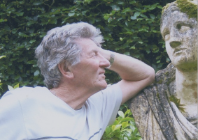 The head and shoulders of a man with grey hair smiling in profile, his left arm resting on the shoulder of an old marble sculpture of a woman whose eyes he is gazing into